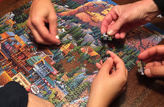 Multiple hands work on a jigsaw puzzle of a forest town.