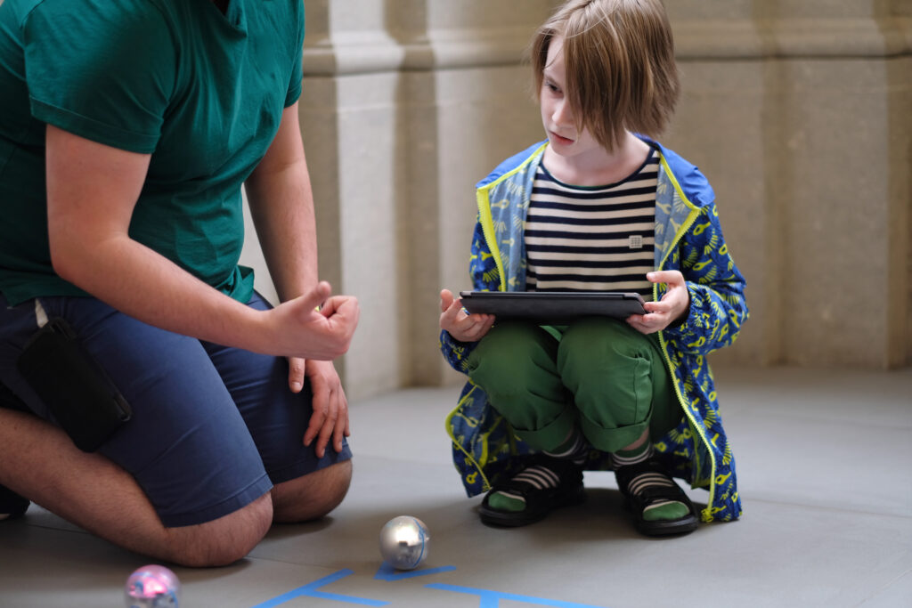 A man explains to a boy the rules of a game with robots controlled from a tablet.