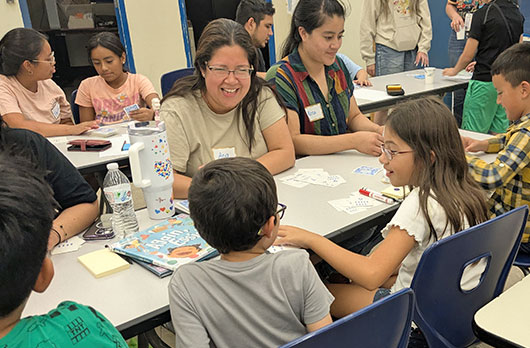 Parents and children sit together at a classroom table, smiling and working on a hands-on math card game activity.