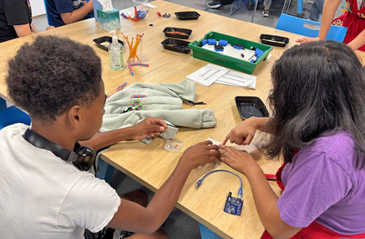 Two students collaborate at a table assembling electronic components connected to a small microcontroller during a NeuroVivid Pathways hands-on STEM activity.