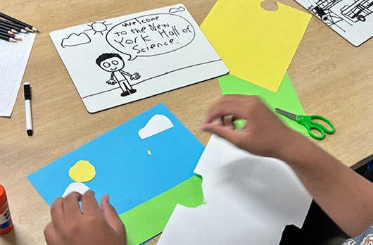 Student creates a paper collage with colored construction paper, scissors, and markers, including a drawing that reads “Welcome to the New York Hall of Science,” during a NeuroVivid Pathways creative STEM activity.