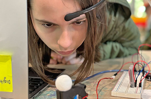 Student wearing a NeuroVivid Pathways brain-computer interface headset concentrates while working with a breadboard and wired electronic components connected to a laptop.