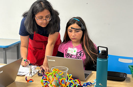 Instructor supports a student using a headband brain-computer interface to control a laptop connected to colorful circuitry during a NeuroVivid Pathways classroom session.